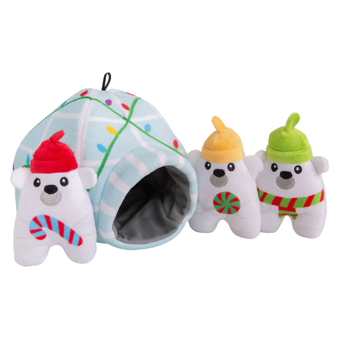 The Snugarooz Hide & Seek Igloo features three plush polar bears in festive hats and scarves, standing before a pastel blue igloo with Christmas lights. Hide treats inside for interactive, holiday fun with your pup!.